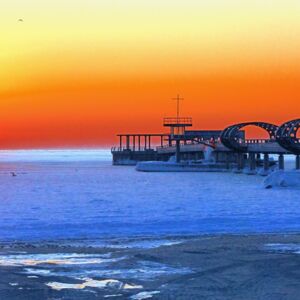 Die Kellenhusener Seebrücke im winter. Am Strand liegt Schnee und am Horizont, der orange leuchtet, geht die Sonne auf