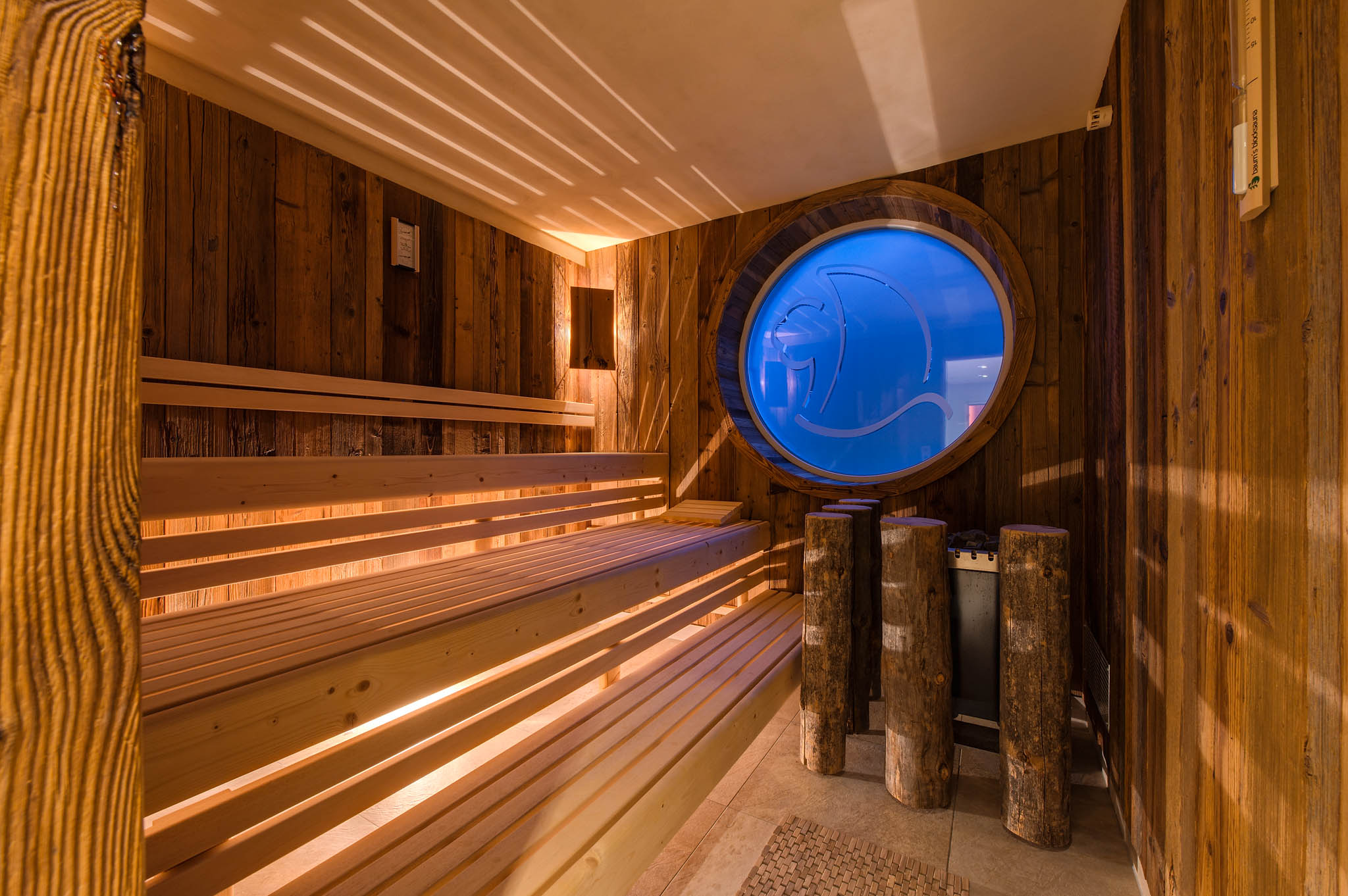 Das Bild zeigt die Saune im Wellness-Bereich vom Hotel ERholung. Man sieht viel Holz und im Hointergrund ein Ficht durchsichtiges Fenster mit dem Logo des Hotels.