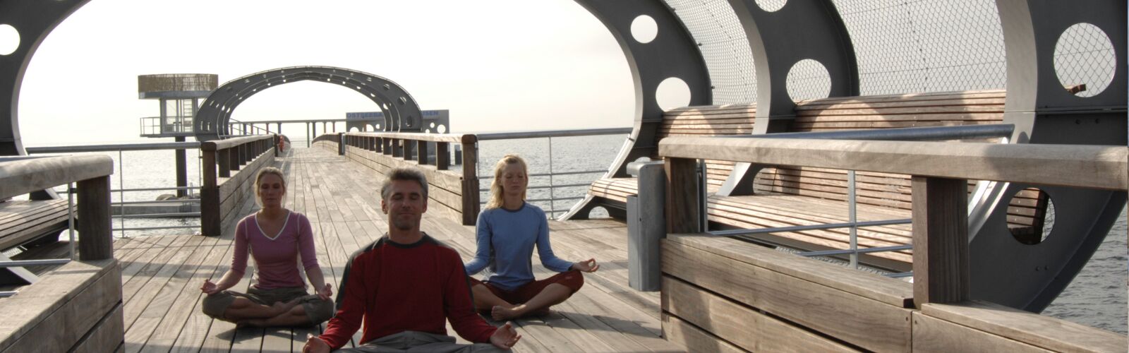 Man sieht einen Mann und zwei Frauen die auf der Kellenhusener Seebrücke sitzen, im typischen Yoga Sitz.